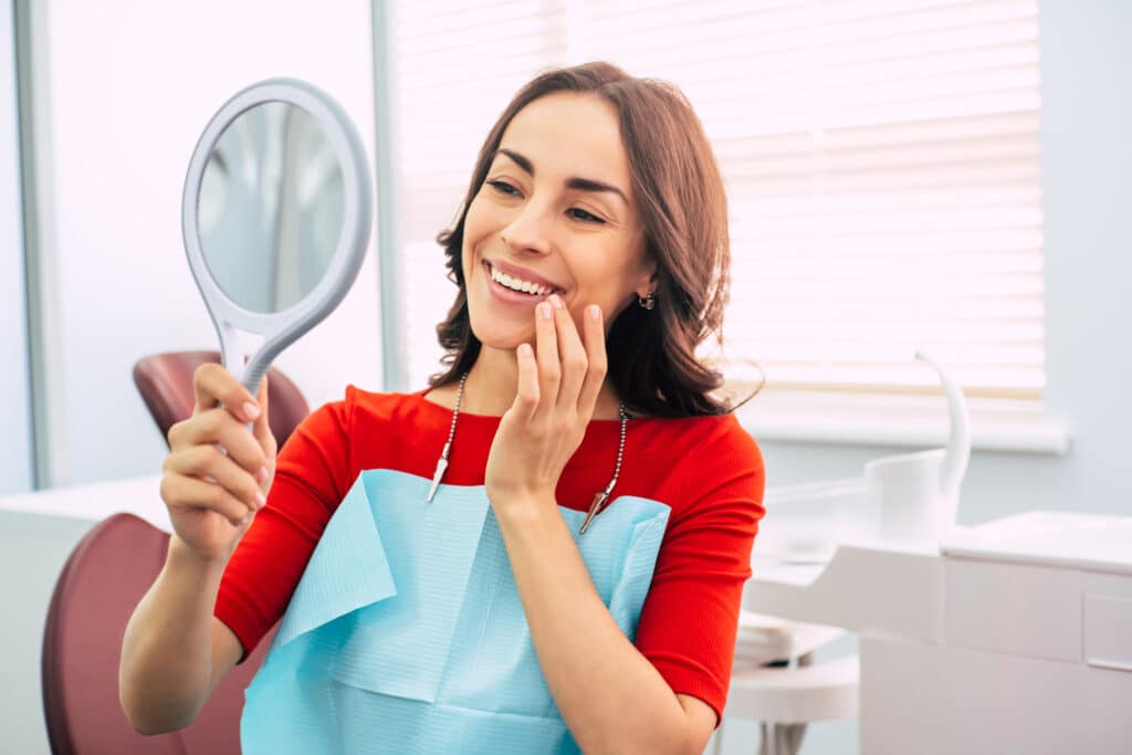Woman in dentist chair looks in the mirror smiling at her new long-lasting dental implants